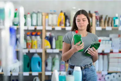 Eine junge Frau steht im Supermarkt vor einem Regal und vergleicht zwei Flaschen Spülmittel.