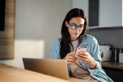 Eine junge Frau mit Brille sitzt an einem Tisch und schaut konzentriert auf ihr Smartphone, während ein Laptop vor ihr steht.