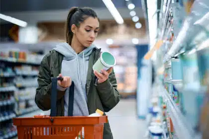 Eine junge Frau steht in einem Supermarkt und studiert konzentriert die Inhaltsstoffe auf der Verpackung eines Produkts, welches seine Rezeptur geändert hat.