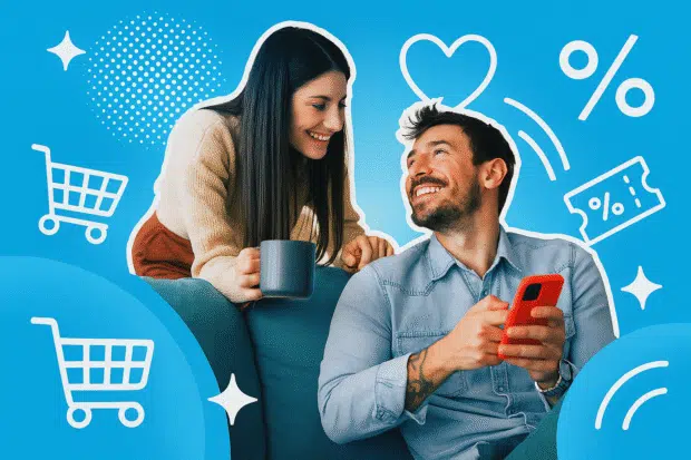 Frau lehnt sich mit Kaffeetasse über die Couch und lächelt ihren Partner an – der ahnt schon, dass sie gleich was Lustiges vorlesen wird.