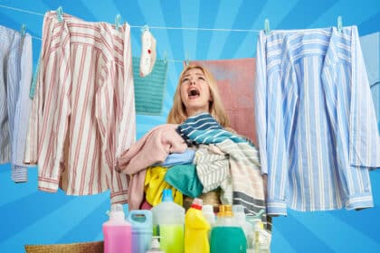 Eine Frau steht zwischen aufgehängter Wäsche, trägt einen großen Haufen bunter Kleidung in den Armen und schaut genervt oder überfordert nach oben vor lauter Haushalt. Vor ihr stehen mehrere bunte Waschmittel-Flaschen.