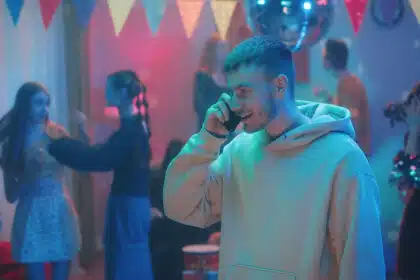 Ein junger Mann in einem Hoodie telefoniert und lächelt, während im Hintergrund bei bunter Partybeleuchtung andere Leute tanzen und sich unterhalten. Bunte Wimpel und eine Discokugel hängen an der Decke – die Szene wirkt wie eine ausgelassene Hausparty.