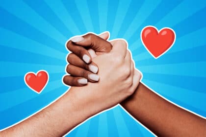 Zwei Hände unterschiedlicher Hautfarben halten sich fest vor einem blauen Hintergrund mit Strahlenmuster; daneben sind zwei rote Herzen abgebildet – ein Symbol für Freundschaft, Zusammenhalt und Liebe.
