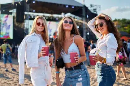 Drei junge Frauen posieren fröhlich mit Getränken in der Hand auf einem Open-Air-Konzert oder Festival. Sie tragen stylische Sommeroutfits und Sonnenbrillen – eine ausgelassene Stimmung, die an Fans bei einem Taylor-Swift-Konzert erinnert.