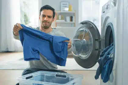 Ein Mann sitzt vor einer Waschmaschine und hält ein blaues Kleidungsstück in den Händen, während er skeptisch oder genervt schaut – möglicherweise ist beim Waschen oder Trocknen etwas schiefgelaufen.