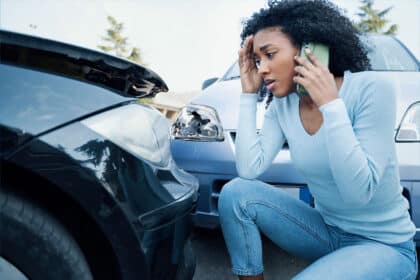Eine Frau hockt besorgt neben zwei Autos mit sichtbaren Unfallschäden und telefoniert, während sie sich mit der Hand an die Stirn fasst.