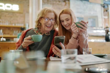 Eine ältere Frau und eine junge Frau sitzen gemeinsam in einem Café, lachen und schauen auf ein Smartphone, während sie Kaffeetassen in der Hand halten.