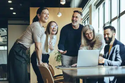 Eine Gruppe von fünf Personen steht und sitzt lachend um einen Tisch mit einem Laptop in einem modernen Büro und scheint gemeinsam etwas Lustiges am Bildschirm zu betrachten.