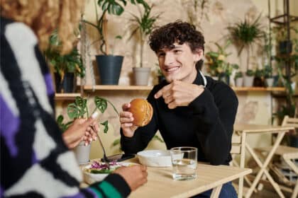 Ein lächelnder junger Mann mit lockigen Haaren präsentiert stolz einen Burger auf einem Holztisch, während er einer Frau gegenüber sitzt, die eine grüne Mahlzeit genießt. Im Hintergrund sind Pflanzen zu sehen.