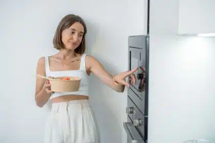 Junge Frau ist an die Wand gelehnt mit ihrem Essen in der Hand und schaltet die Mikrowelle aus.