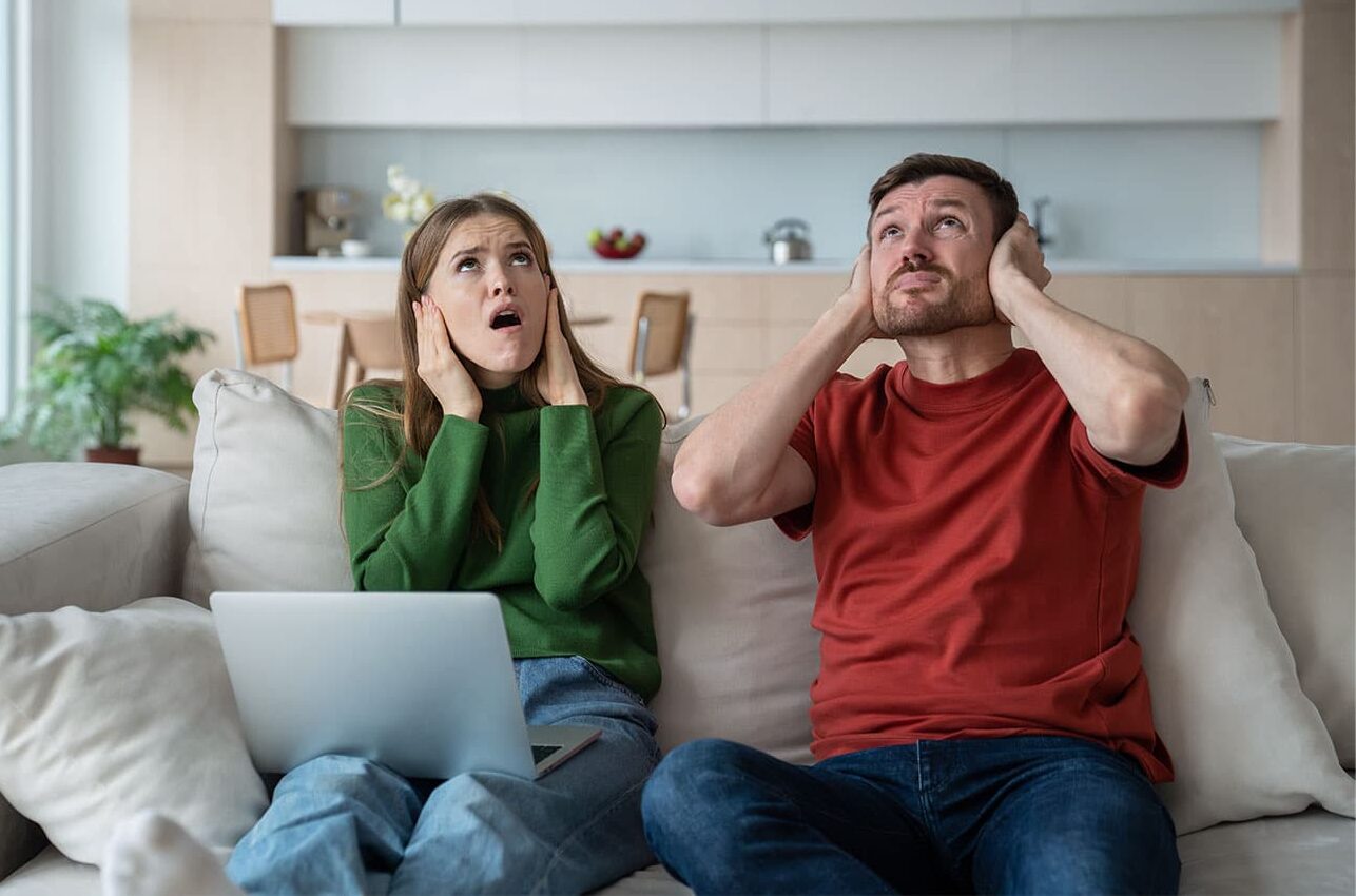 Ein Mann und eine Frau sitzen auf einem Sofa in einem Wohnzimmer. Beide halten sich genervt die Ohren zu und blicken nach oben, als würden sie von lautem Lärm aus der Wohnung über ihnen gestört.