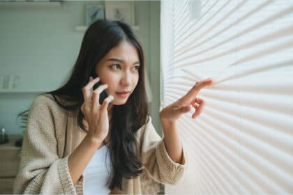 Eine junge Frau blickt vorsichtig durch geschlossene Jalousien nach draußen, während sie mit einem Smartphone telefoniert.