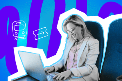 Frau arbeitet lächelnd an einem Laptop im Zug; sie sitzt auf einem blauen Sitzplatz.