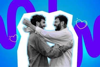 Zwei Männer umarmen sich lachend und blicken sich liebevoll an, vor einem farbigen Hintergrund mit Herzsymbolen.